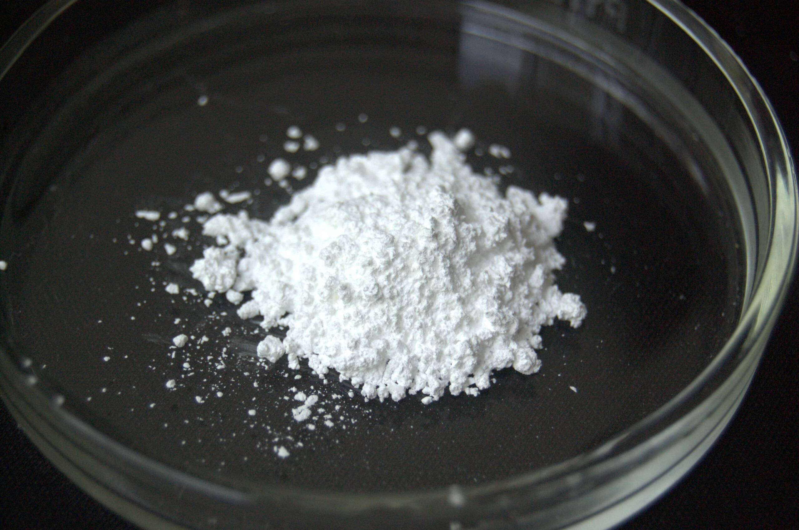 A pile of indium hydroxide sits as a white powder on a clear glass petri dish against a stark black background.