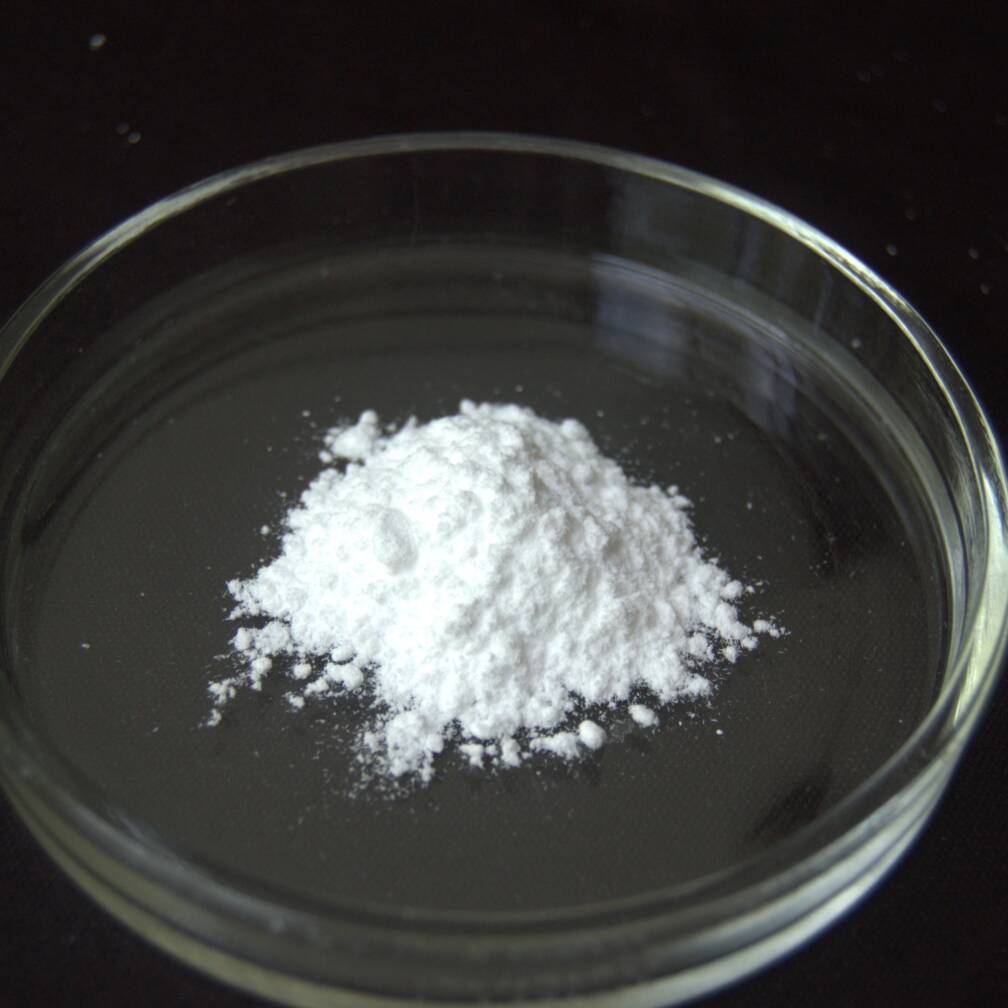 Một loại bột màu trắng của indium acetate nằm trong một đĩa petri thủy tinh trong suốt trên nền tối.