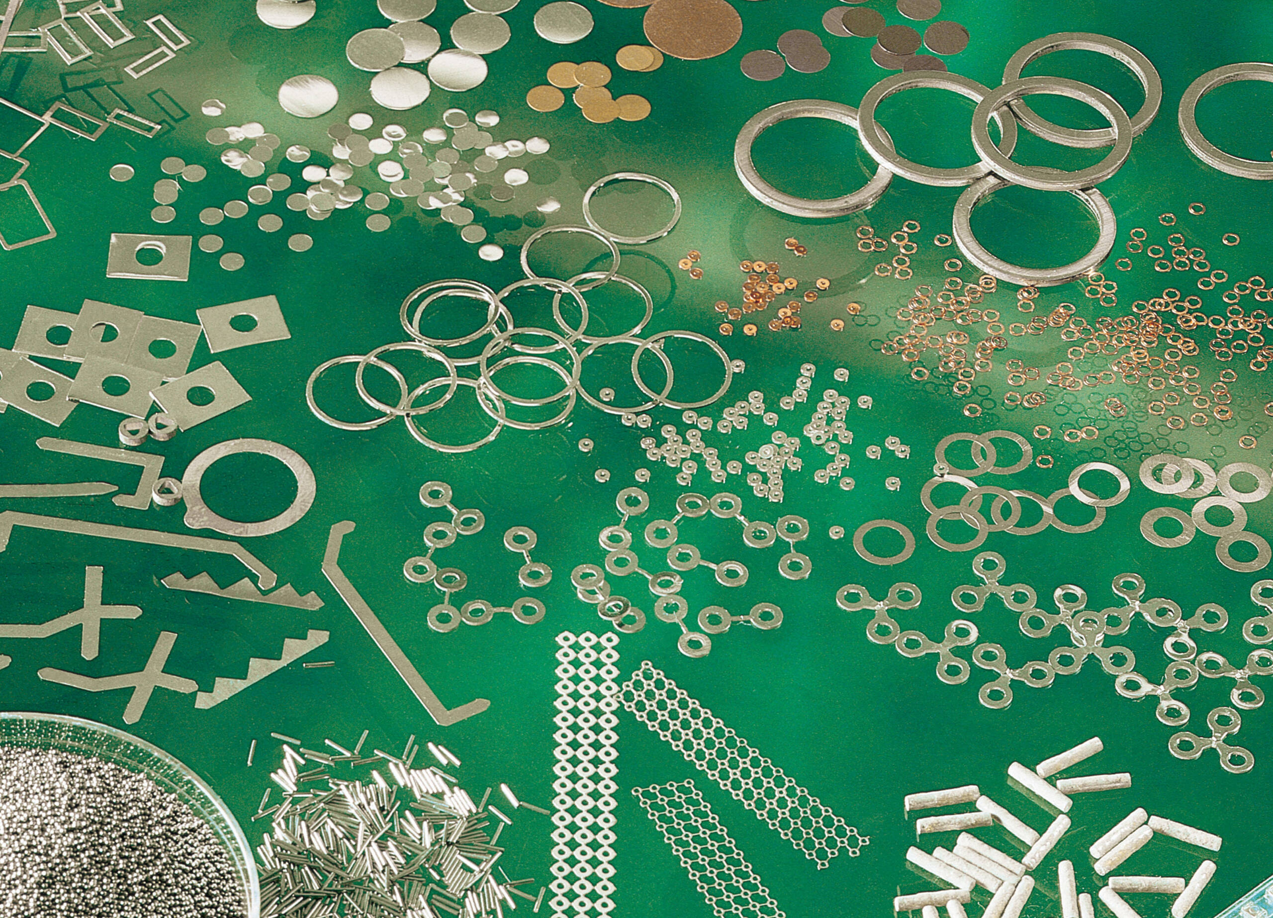 Flux coated solder preforms and assorted small metal components, including rings, discs, rods, and wires, are meticulously arranged on a green surface.