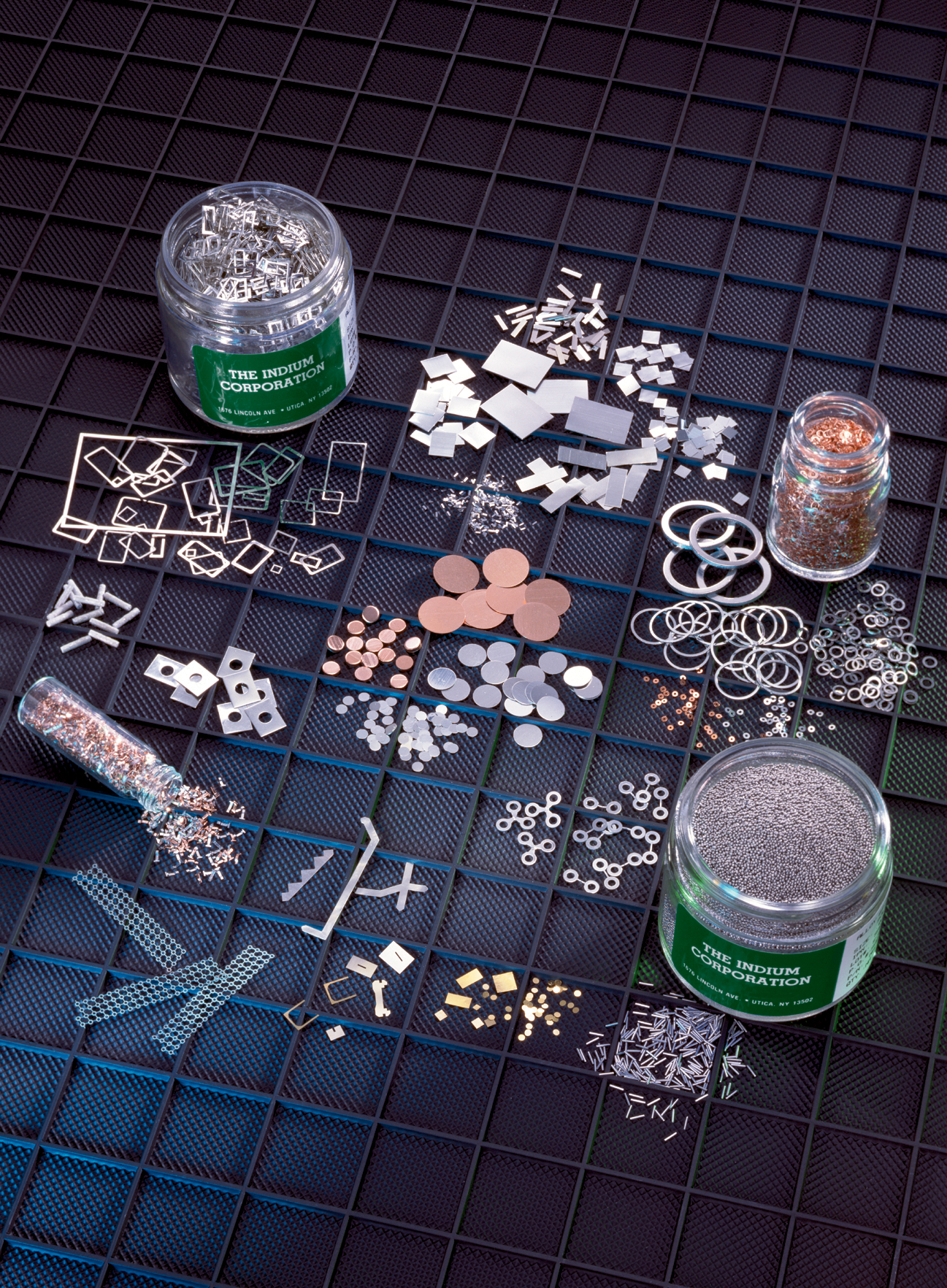 Various metal shapes, small components, and solder preform 140s are scattered around glass jars on a grid-patterned surface.