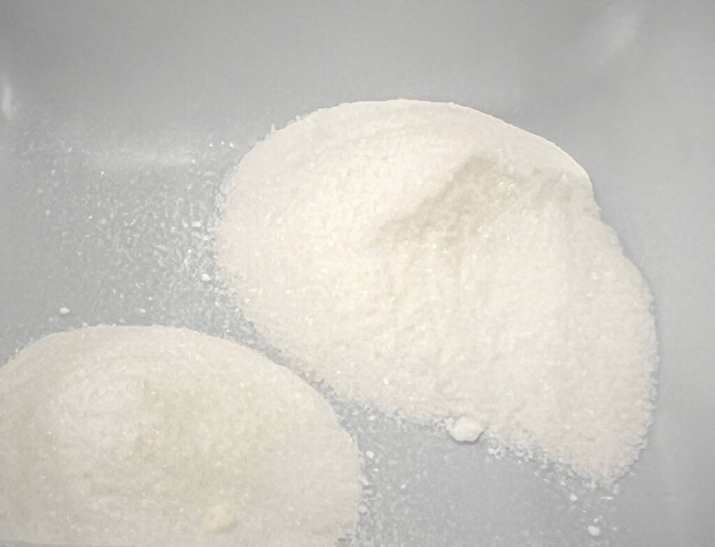 Auf einer glatten, hellen Oberfläche befinden sich zwei Häufchen mit weißem Pulver, möglicherweise Acetylacetonat.