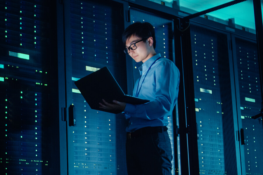 In a dimly lit server room, a person works with precision on their laptop amidst glowing blue servers, resembling the intricate art of computer solder.