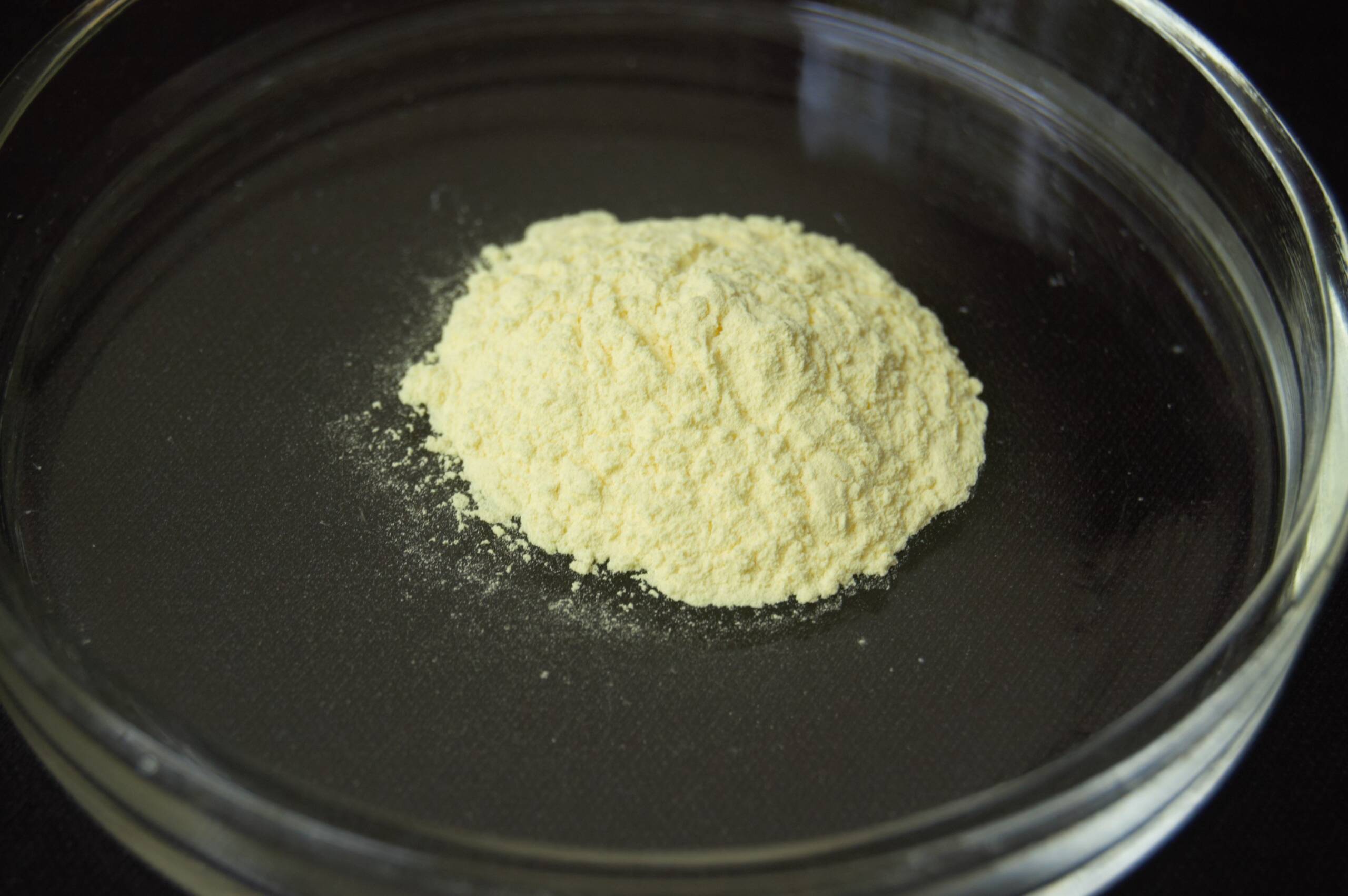 A mound of yellow indium tin oxide powder sits in a clear glass petri dish on a black background.