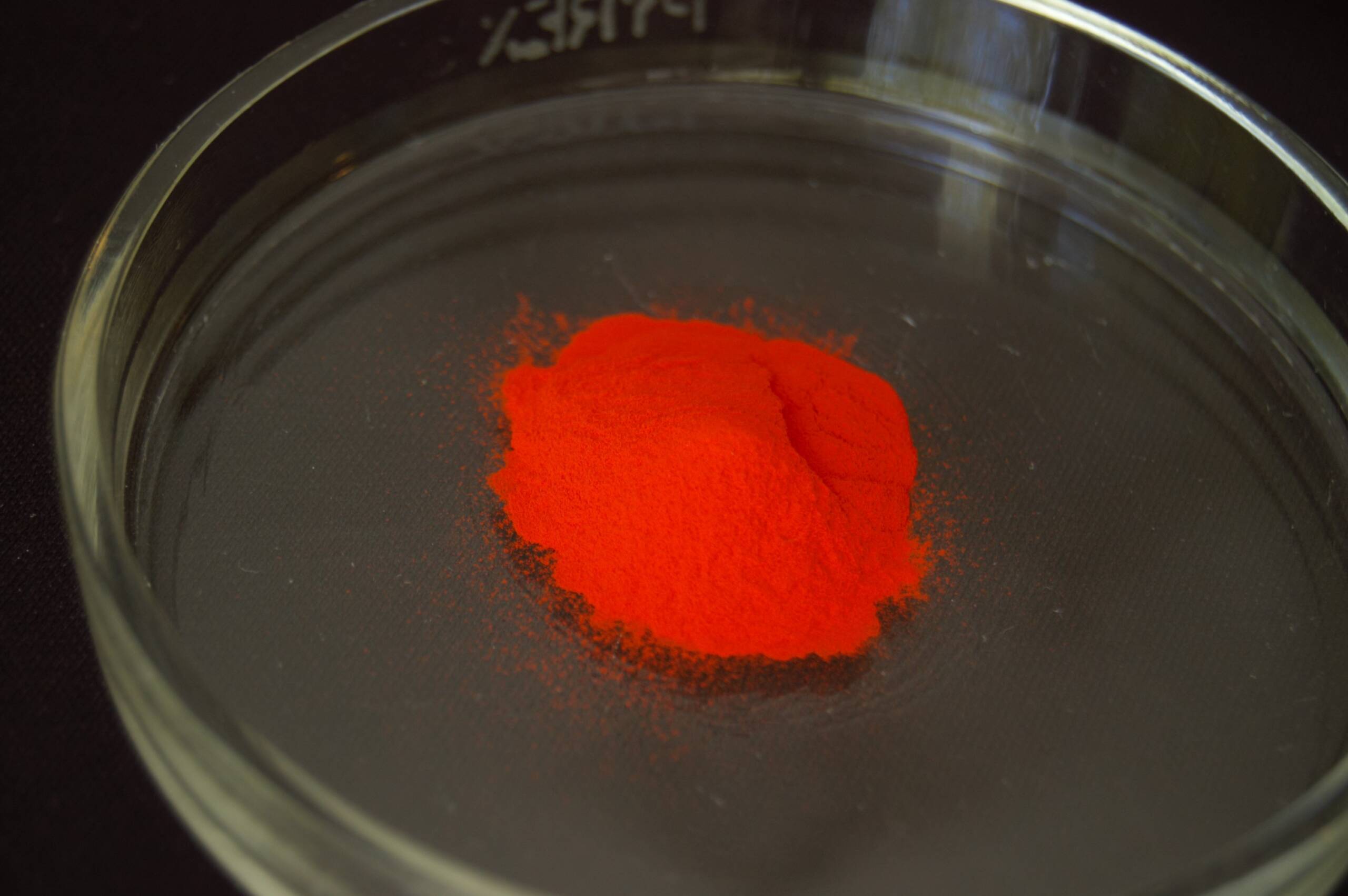 Bright orange indium sulfide powder rests in a clear glass dish against a dark background.