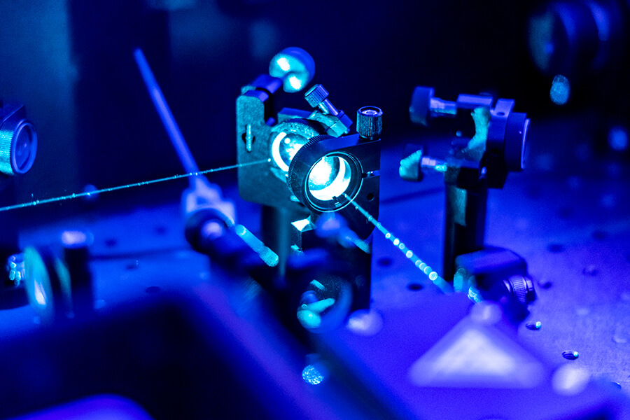 A bluish laboratory setup features fiber optic lasers focusing through lenses and optical components on a metal surface.