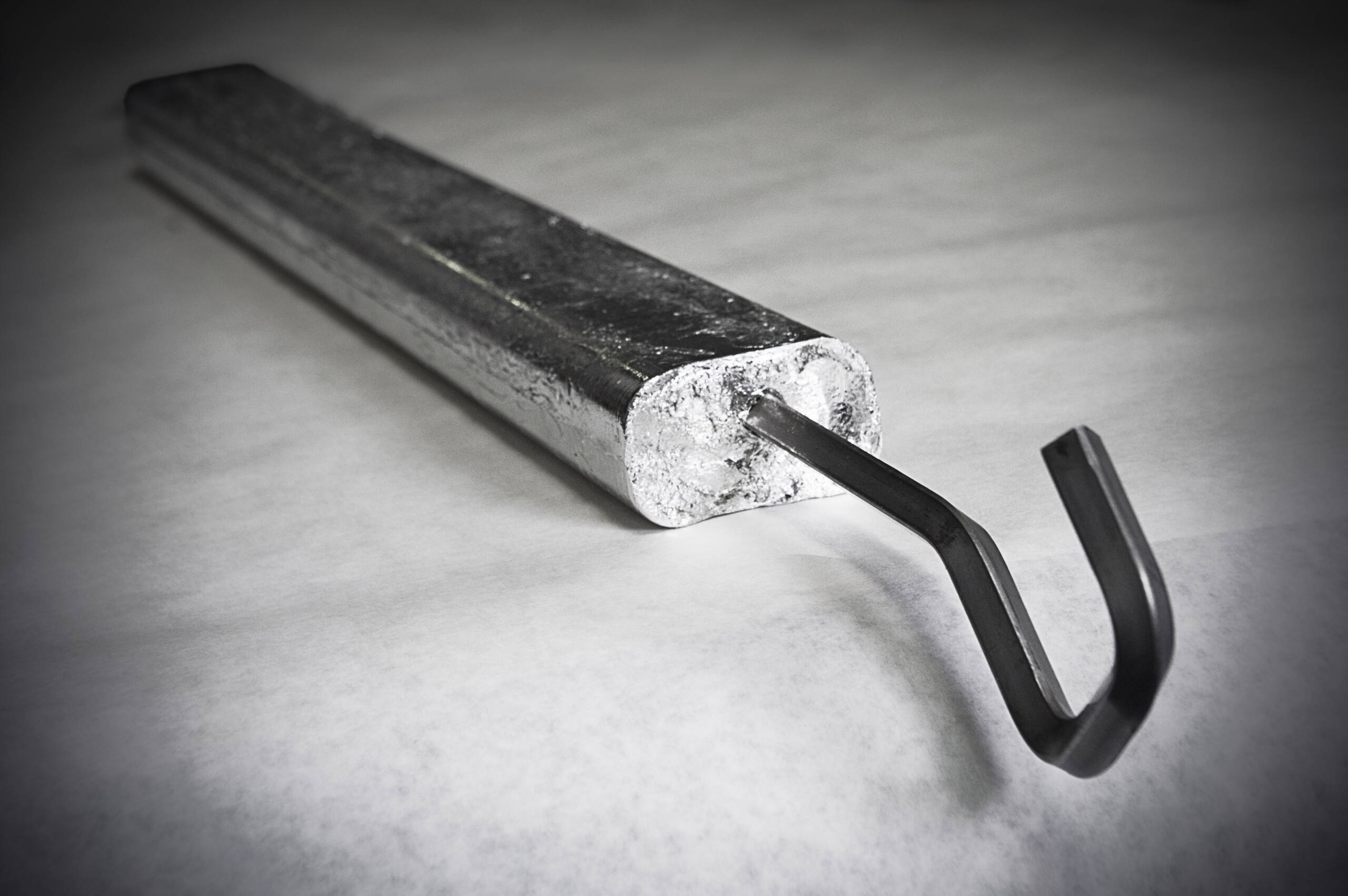 Rectangular metal rod with a curved hook at one end, resembling an indium electroplating anode, placed on a light surface.