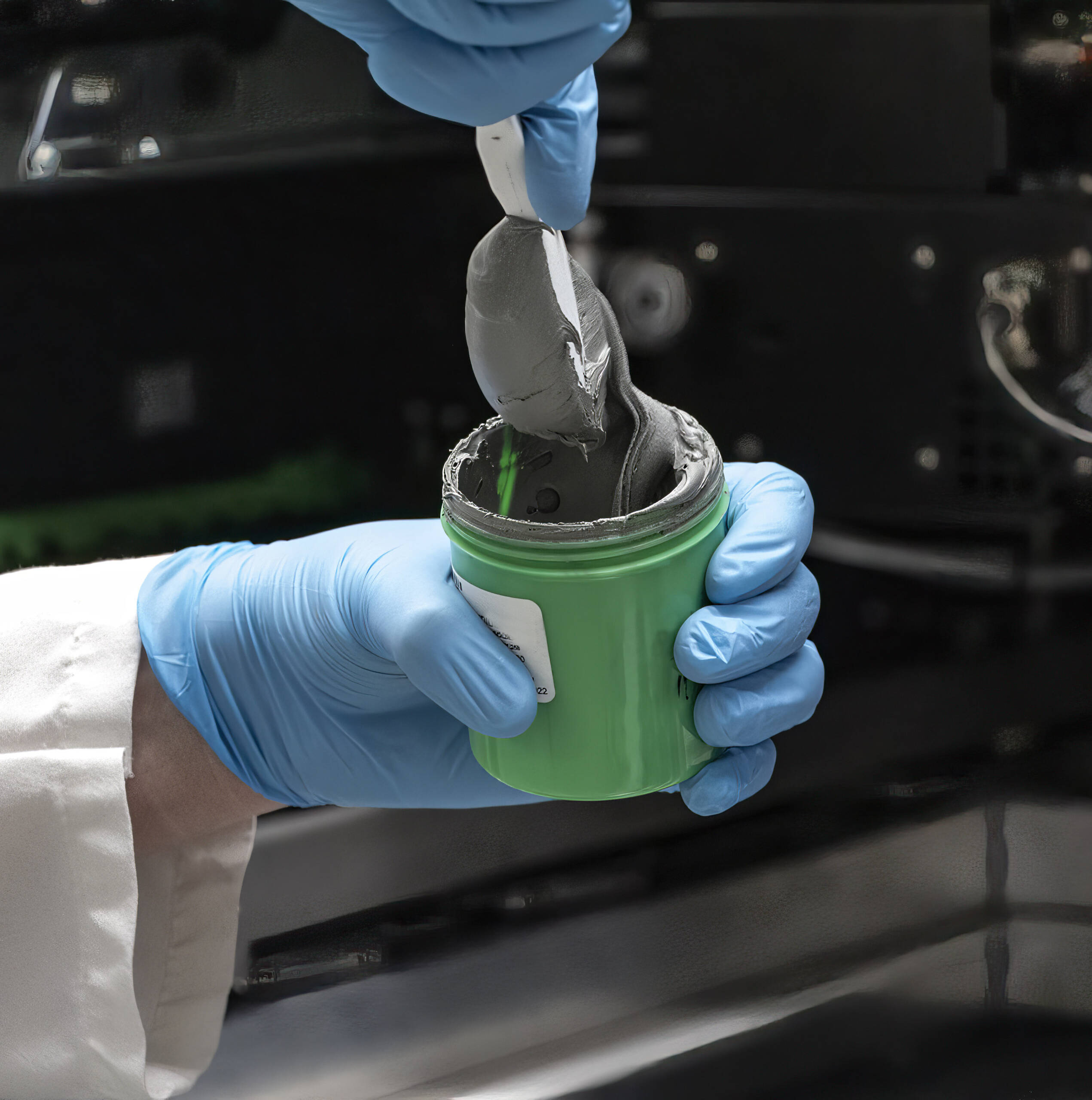 A gloved hand scoops gray, Pb-free SAC paste from a green container with a spatula in a laboratory setting.