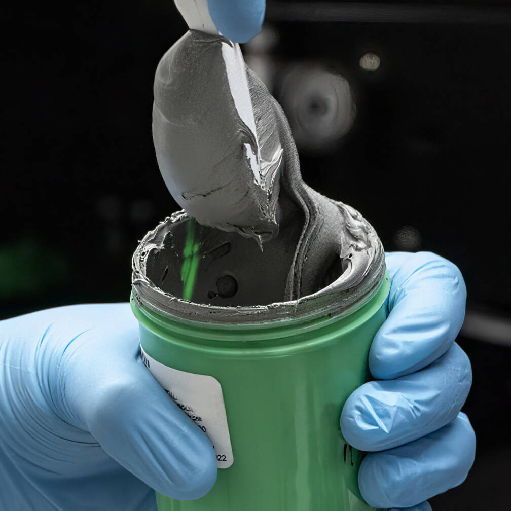 A gloved hand scoops thick gray lead free solder paste from a green container with a metal spatula.