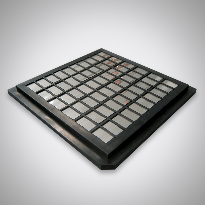 A black, square plastic grid cover with a raised edge, placed on a light gray surface beside a gold alloy solder preform.