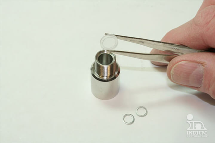 A hand uses tweezers to place a small round object into a metal holder, with two similar cryogenic seals nearby.