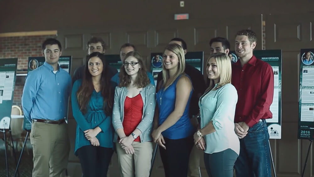 A group of cheerful interns stands smiling in front of academic posters, capturing the moment for the archive.