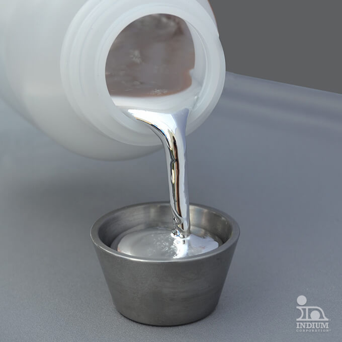 Eine Flasche gießt ein flüssiges Metall in einen kleinen Metallbecher auf einer grauen Fläche.