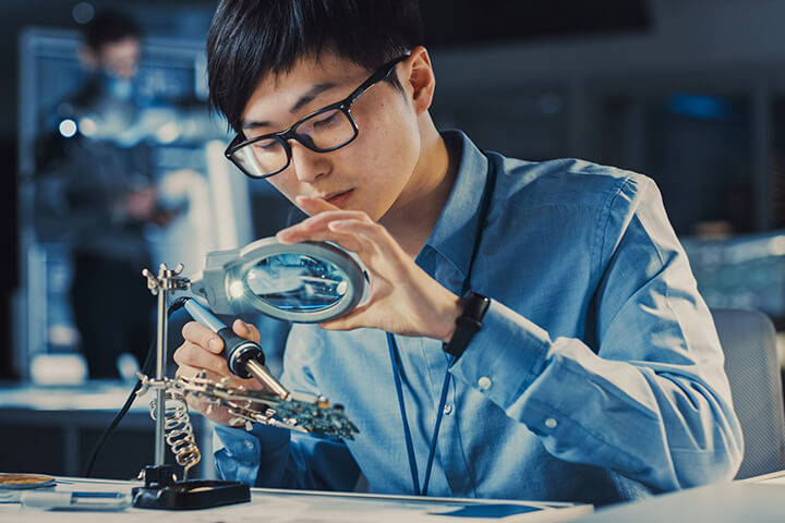 Male engineer soldering electronics while looking through magnifying glass.
