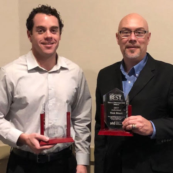 Two men in business attire are standing in the Media Center, proudly holding awards and smiling at the camera.