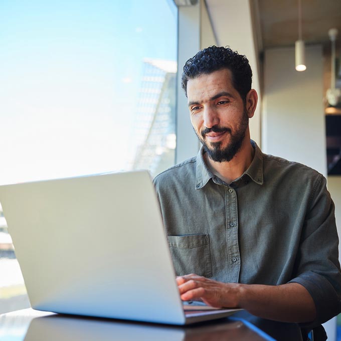 Curly-haired man with a beard works on his laptop by a window seeking sales support.