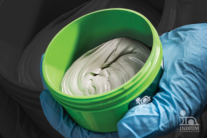 A gloved hand holding an open green container of solder paste