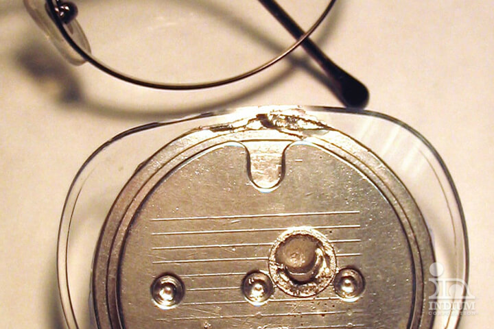 Close-up of a metallic cochlear implant device with eyeglasses in the background on a light surface.