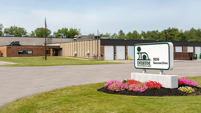 Sign for Indium Corporation at 5366 Success Drive, in front of a large industrial building with a lawn and flowers.