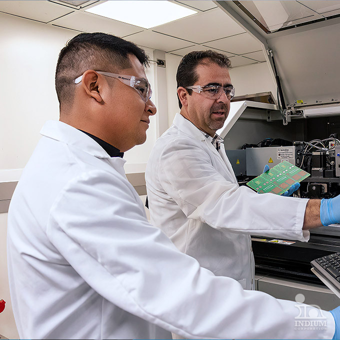 Two scientists in lab coats and safety glasses examine a circuit board in a laboratory setting.