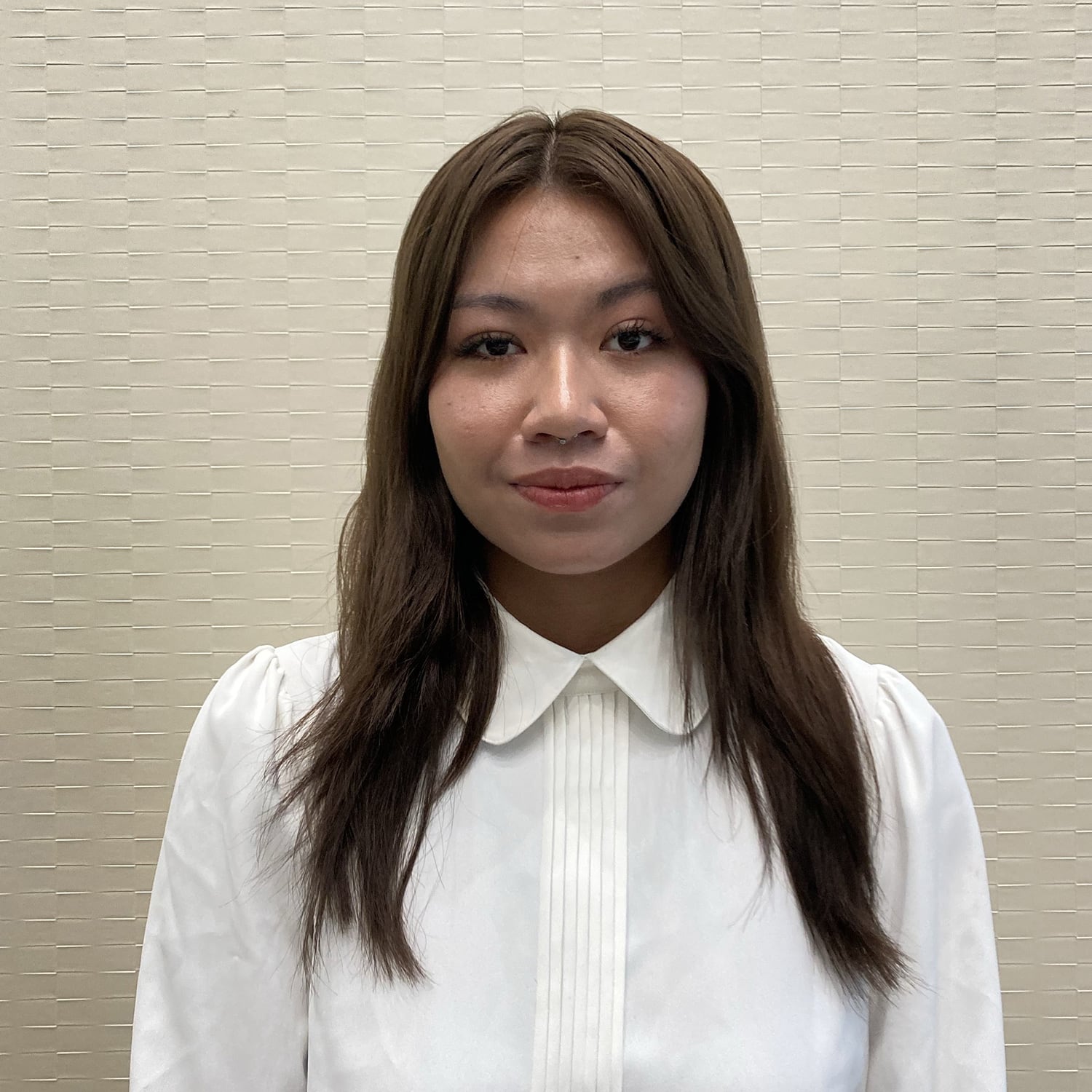A woman with long brown hair in a white shirt stands against a textured beige wall, exuding calm amidst the bustling world of current interns.