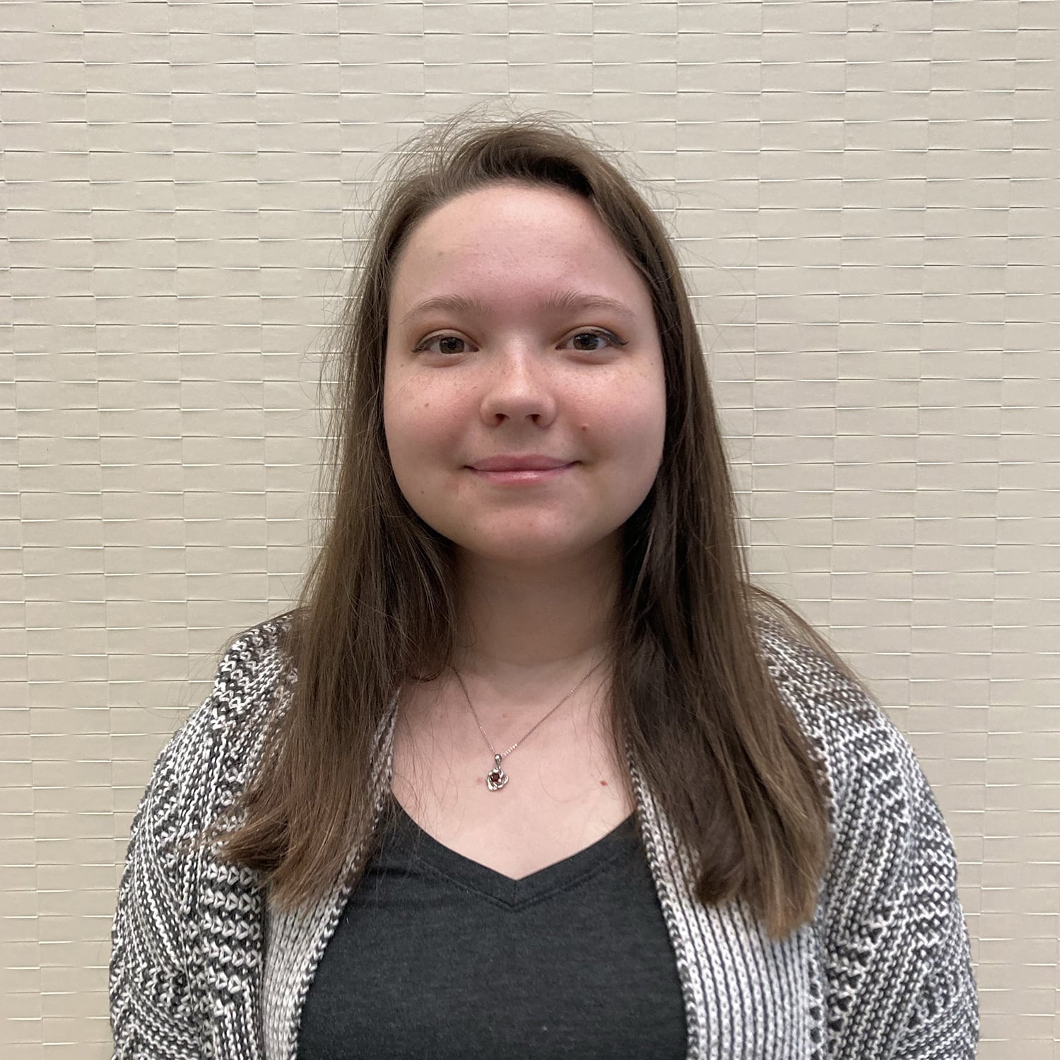 Young woman with long brown hair and a patterned cardigan, smiling slightly in front of a light textured wall, embodies the vibrant spirit of our current interns.