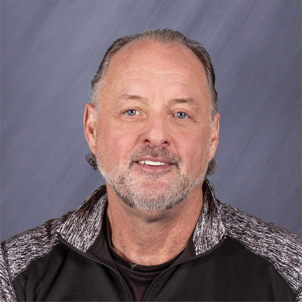 Mike McKenna, a man with short gray hair and a beard, smiles warmly in a portrait while wearing his black and gray shirt.